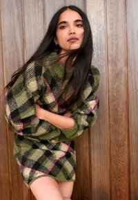 Green wool sweater dress features a checkered pattern with pink accents. It has a relaxed fit and wide sleeves, set against a wooden background.
