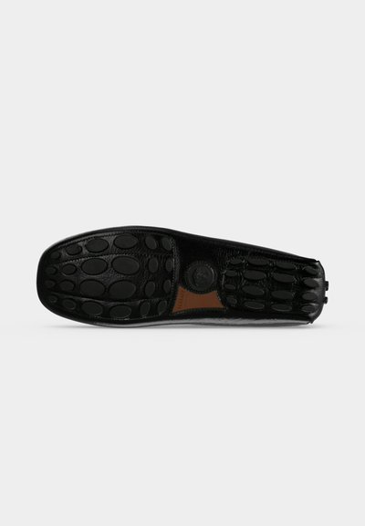 Semelle de mocassin en cuir noir avec des grips en caoutchouc ovales et un petit patch en cuir marron au centre, présenté sur un fond clair uni.