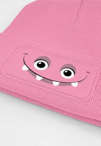 Pink knitted beanie featuring a rectangular patch with a smiling monster face, large eyes, and two protruding teeth. Textured fabric design.