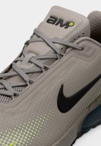 Αθλητικό παπούτσι ανοιχτού γκρι με υφασμάτινο επάνω μέρος, κυκλικές οπές, μαύρο λογότυπο Nike swoosh, γκρι κορδόνια και διακριτική πράσινη λεπτομέρεια.