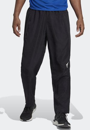 Homme portant un pantalon de sport noir ample avec des poches latérales et des chaussures de course noires, debout devant un fond blanc.