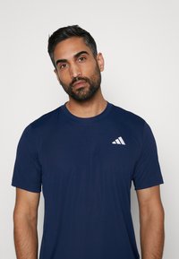 Camiseta deportiva azul marino hecha de tejido transpirable con mangas cortas. Presenta un logo blanco de Adidas en la parte izquierda del pecho.