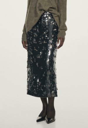 Personne portant une jupe midi foncée à sequins, des collants noirs, des talons aiguilles noirs pointus et un pull marron à manches longues.