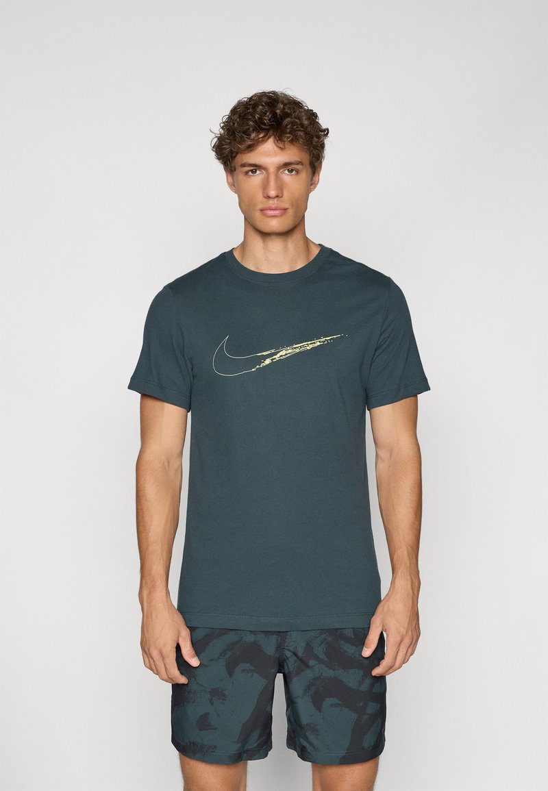 Temnozelena bombažna majica s zlatim logotipom Nike swoosh. Dopolnjena s črnimi kratkimi hlačami s vzorcem in zelenimi detajli.