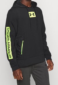 Felpa con cappuccio nera dotata di un grande logo verde neon sul petto e sulla tasca laterale, con il testo in grassetto "UNDER ARMOUR" sulla manica.