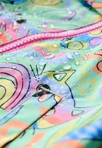 Uimahousukangas pastellisävyissä abstrakteilla kuvioilla, jossa on vaaleanpunainen vetoketju ja pinnalla vesipisaroita, korostaen vedenpitävyyttä.