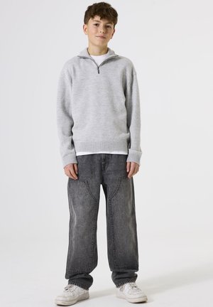 Lichtgrijze sweater met rits en geribbelde zoom; losse, donkergrijze jeans met uniek zakontwerp; witte sneakers.