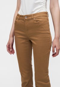 Femme portant un pantalon slim marron taille haute avec poches avant et un haut blanc froncé sur le côté, mains détendues le long du corps.
