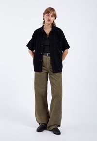 Svart kortärmad skjorta med ficka, bärs över en mörk linne. Olivgröna tuskaff pants med vida ben. Svarta skor med en texturerad yta.