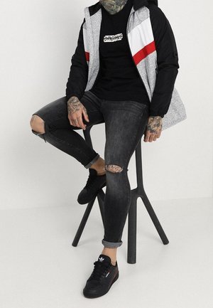 Schwarz-weiß gemusterte Jacke mit roten Akzenten, schwarzes T-Shirt, zerrissene schwarze Jeans und schwarze Sneakers sitzen auf einem Hocker vor einem weißen Hintergrund.