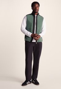 Green fleece vest with black zip and trim, worn over a white long-sleeve shirt. Model in black trousers and dark sneakers.