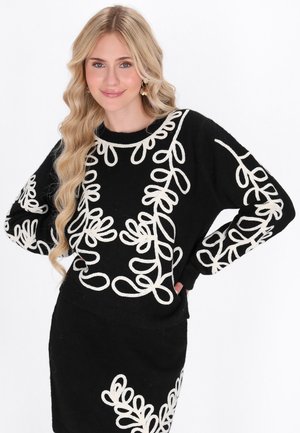 Schwarzer Pullover mit cremefarbener Loop-Stickerei. Lange Ärmel und runder Halsausschnitt. Kombiniert mit einem passenden schwarzen Rock mit ähnlicher Stickerei.