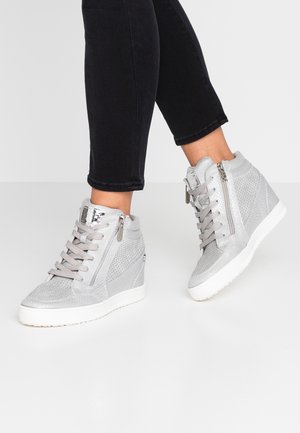 Person iført grå kile-sneakers med snørebånd og lynlåse, kombineret med sorte tætsiddende bukser mod en ensfarvet lys baggrund.