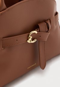 Bruine leren handtas met een gestikte gespband en een goudkleurige "C"-sluiting, voorzien van een klein gouden "Coccinelle"-logo.