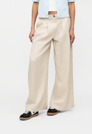 Asmuo dėvi plačias smėlio spalvos kelnes, juodus sportbačius su baltomis raištelėmis ir šviesiai mėlyną trumpą švarkelį ant baltos palaidinės.