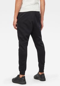 Svarta träningsoveraller i bomullsmix; smalnande design med sidoficka; elastiska muddar; bärs med svarta sneakers.