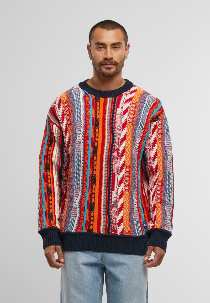 Mann, der einen bunten, gemusterten Strickpullover mit vertikalen Streifen in Rot, Orange, Blau und Weiß trägt, kombiniert mit hellblauen Jeans.