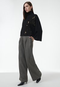 Schwarzes, kurzes Oberteil mit Gittermuster und goldenen Details, kombiniert mit weiten grauen Hosen. Glatte Textur und spitze schwarze Absätze runden den Look ab.