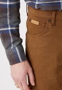 Höger hand med guldring nära frampocket på bruna Wrangler-byxor, i kombination med mudd på blå och brun rutig skjorta med långa ärmar.