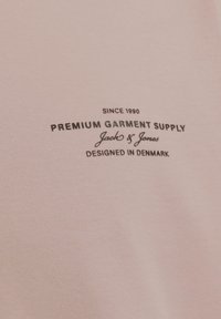 Svetlo roza tkanina z gladko teksturo vsebuje črno natisnjeno besedilo: "PREMIUM GARMENT SUPPLY," "Jack & Jones," in "DESIGNED IN DENMARK."