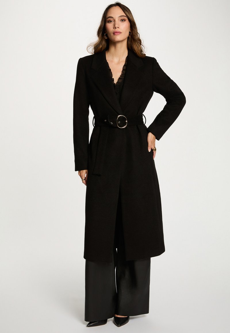 Lang zwarte jas met een riem en ronde gesp, gestructureerde schouders en een open voorkant. Gestyled met brede zwarte broek.