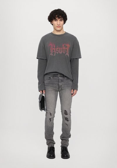 T-shirt graphique surdimensionnée grise avec un texte en flammes rouges, portée avec un jean gris déchiré et des bottes noires à lacets. Tenant un sac noir.