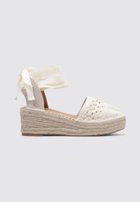 Cuña blanca de espadrille con parte superior de tela trenzada, recortes decorativos y lazos de cinta en el tobillo. La suela envuelta en yute natural añade textura.