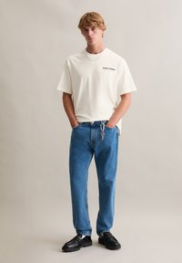 Wit t-shirt met korte mouwen en de tekst "EARLY BIRDS", gecombineerd met blauwe spijkerbroek en zwarte opvallende schoenen. Eenvoudig ontwerp en een casual pasvorm.