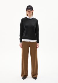 Schwarzer Pullover mit Rundhalsausschnitt, über einem weißen Hemd mit Kragen getragen, kombiniert mit braunen weiten Hosen und einer schwarzen Baseballkappe.