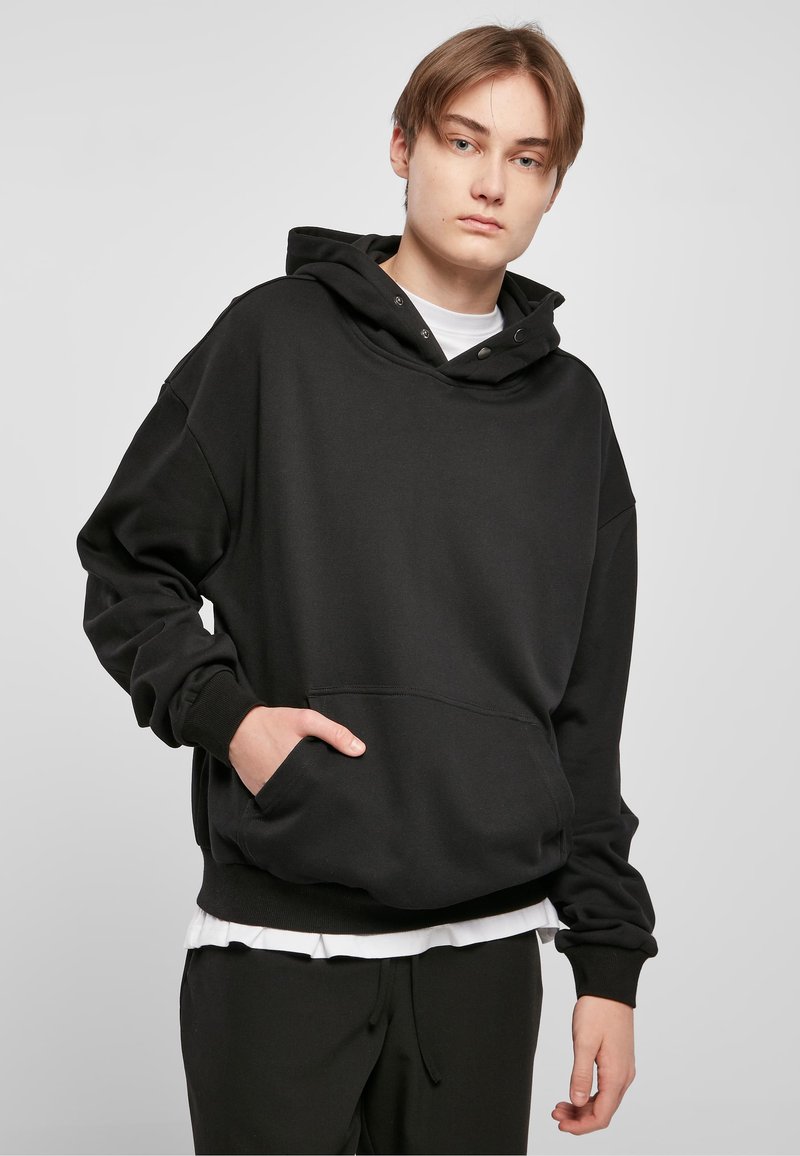 Black oversized hoodie made of soft fabric, featuring a kangaroo pocket, drawstring hood, and ribbed cuffs, paired with a white undershirt.