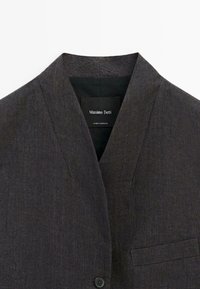 Mörkgrå kavaj med slagkrage, tillverkad av textil med struktur. Har en enkelknappsstängning och en bröstficka. Etikett synlig.