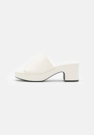 Witte open teen mule sandaal met gestructureerd bovenwerk, blokhak en plateauzool op een effen witte achtergrond.