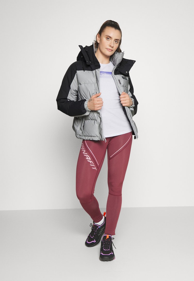 Grau und schwarze Steppjacke, eng anliegende burgunderfarbene Leggings mit weißen Akzenten, schwarze Sneakers mit bunten Details, weißes T-Shirt darunter.