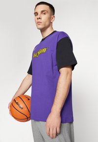 Tricou scurt purpuriu cu detalii negre și inscripția „Lakeshow” imprimată în galben. Modelul ține o minge de baschet și poartă pantaloni de trening gri.