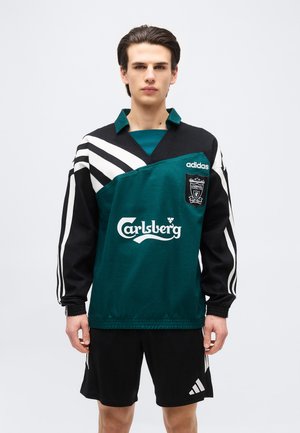 Junger Mann trägt ein grünes und schwarzes Adidas Liverpool Fußballtrikot mit Carlsberg Sponsorenlogo und schwarze Adidas-Shorts.