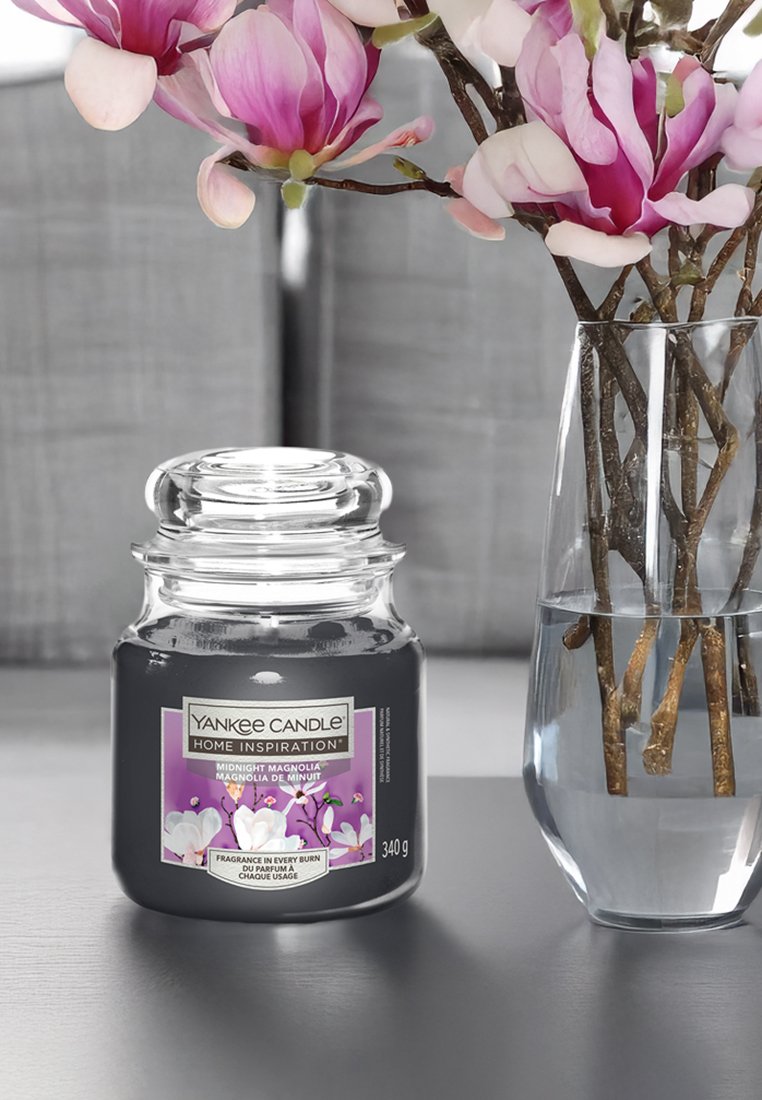 Vaso Yankee Candle con coperchio in vetro trasparente, base nera e etichetta viola con fiori di magnolia. Circondato da un vaso trasparente con rami.