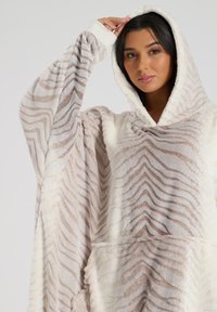 Hyggeligt oversized hætte tæppe, blødt stof i cremede og beige zebra mønster, har lange ærmer og en frontlomme for varme og komfort.