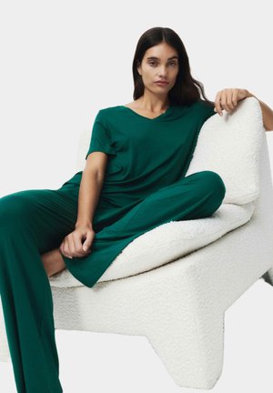 Conjunto de salón verde en tela suave con diseño en forma de V. Sentada en una silla escultórica blanca y texturizada. Corte relajado, mangas largas y piernas anchas.