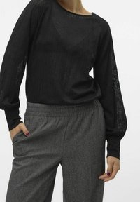 Kvinna som bär en genomskinlig svart blus med långa ärmar, instoppad i högmidjade grå vida byxor med elastisk midja, med händerna placerade nära fickorna.