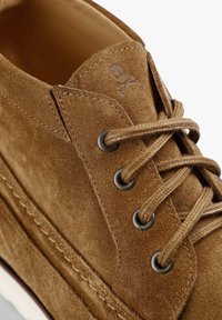 Chaussure en daim marron avec des œillets métalliques ronds, des lacets assortis et un petit crâne et os croisés embossés près de la languette.