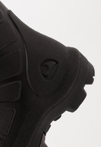 Shoe atlético preto com superfície texturizada, padrões elevados e lugs de sola distintos; apresenta o logotipo em relevo na lateral.