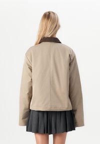 Veste beige courte avec un col en velours côtelé marron, dotée de manches longues et d'une texture lisse. Assortie avec une jupe noire plissée.