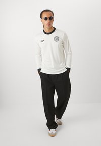 Fehér, hosszú ujjú ing, texturált anyagból, fekete nyakkal és Adidas logóval. Fekete nadrággal és piros részletekkel ellátott fehér sportcipővel párosítva.