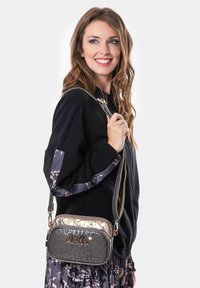 Mujer con cabello largo y ondulado sonríe, vistiendo una chaqueta negra y una falda estampada, sosteniendo un pequeño bolso cruzado con el logo "Anekke".
