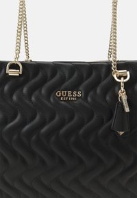 Borsa a tracolla nera trapuntata con una tracolla in catena dorata, logo in rilievo "GUESS EST 1981" e un piccolo tag a forma di portachiavi. Presenta una texture liscia e un motivo ondulato.