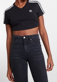 Černé zkrácené tričko s výstřihem do V, na ramenou zdobené třemi bílými pruhy a malým bílým logem. V kombinaci s tmavými džínami s vysokým pasem.