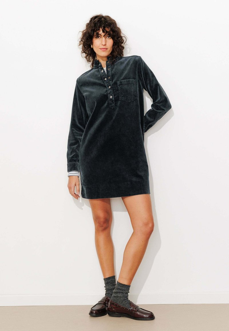 Robe en velours côtelé vert foncé avec des manches longues, patte de boutonnage et poche poitrine. Assortie à des chaussettes grises et des mocassins marron.