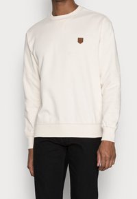 Krémová mikina s dlouhými rukávy, na manžetách a dolním lemu s žebrovaným vzorem, s malou hnědou koženou nášivkou na hrudi. Jednoduchý design a textura.