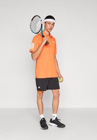 Camiseta de tenis naranja con tres rayas blancas en la manga izquierda, combinada con pantalones cortos negros, sosteniendo una raqueta y una pelota de tenis, llevando zapatillas deportivas.