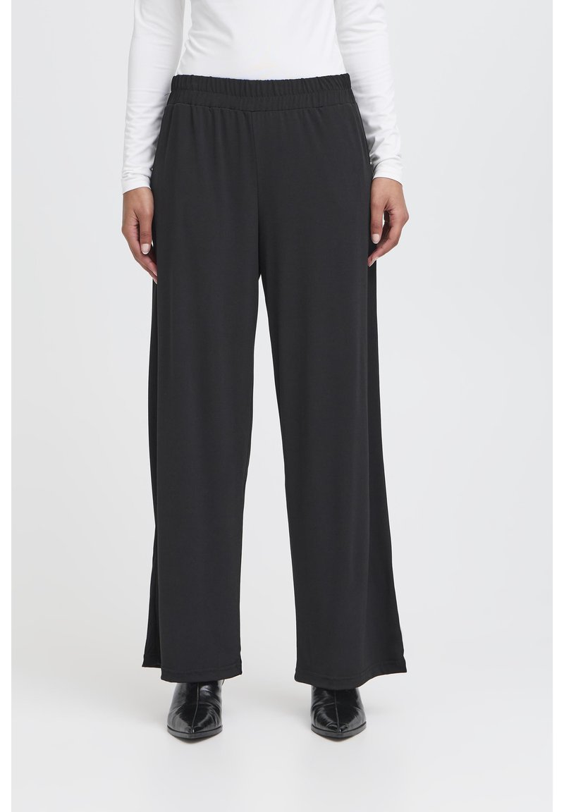 Femme portant un pantalon large noir avec une taille élastique et des bottines noires à bout pointu, un haut blanc à manches longues partiellement visible.